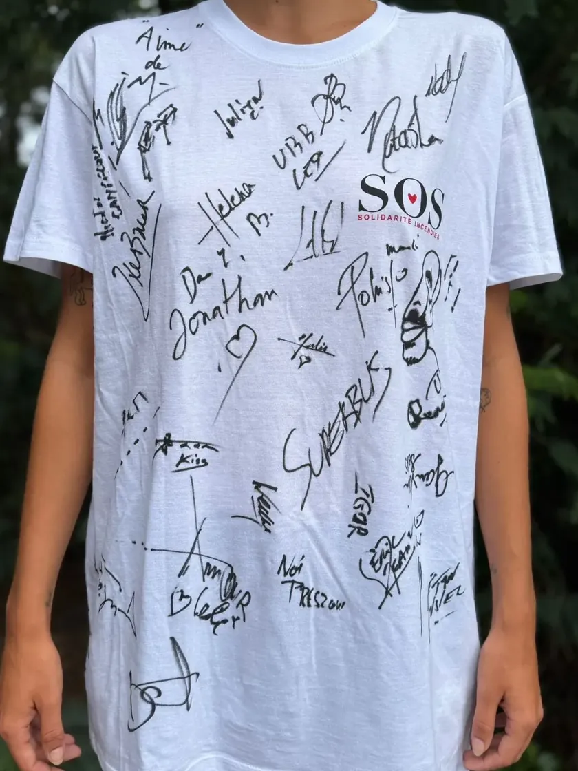 Recto du t-shirt blanc de la tombola, couvert des signatures manuscrites des artistes du concert, avec le petit logo SOS Solidarité Incendies sur la poitrine.